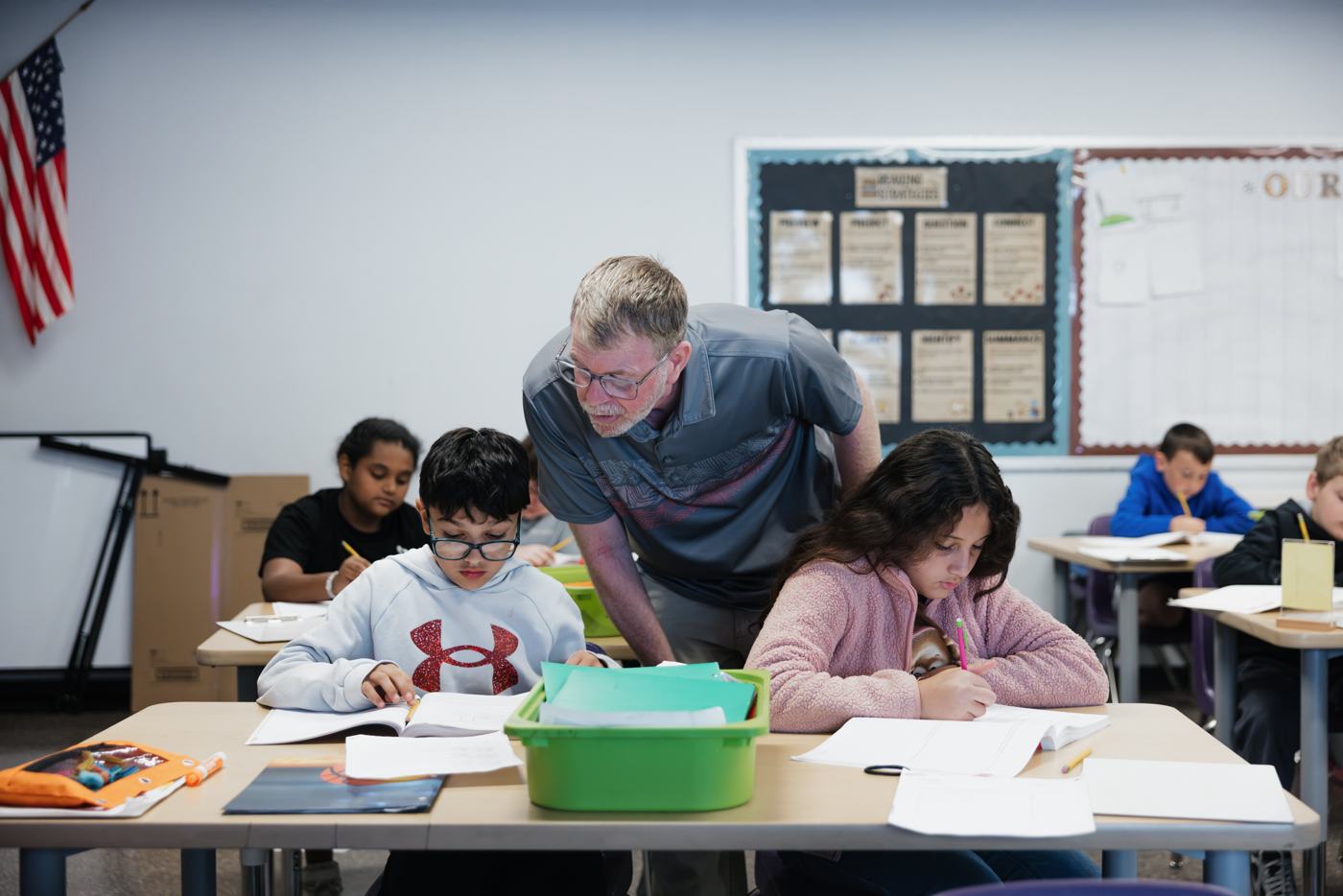 A Marion teacher leans in to coach two students as they read and write at their desks.