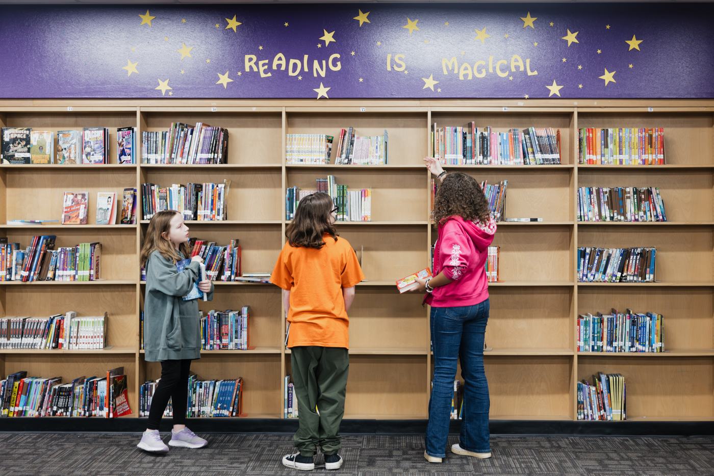Three Marion students browse library shelves under a purple “Reading is Magical” banner, one reaching for a book.