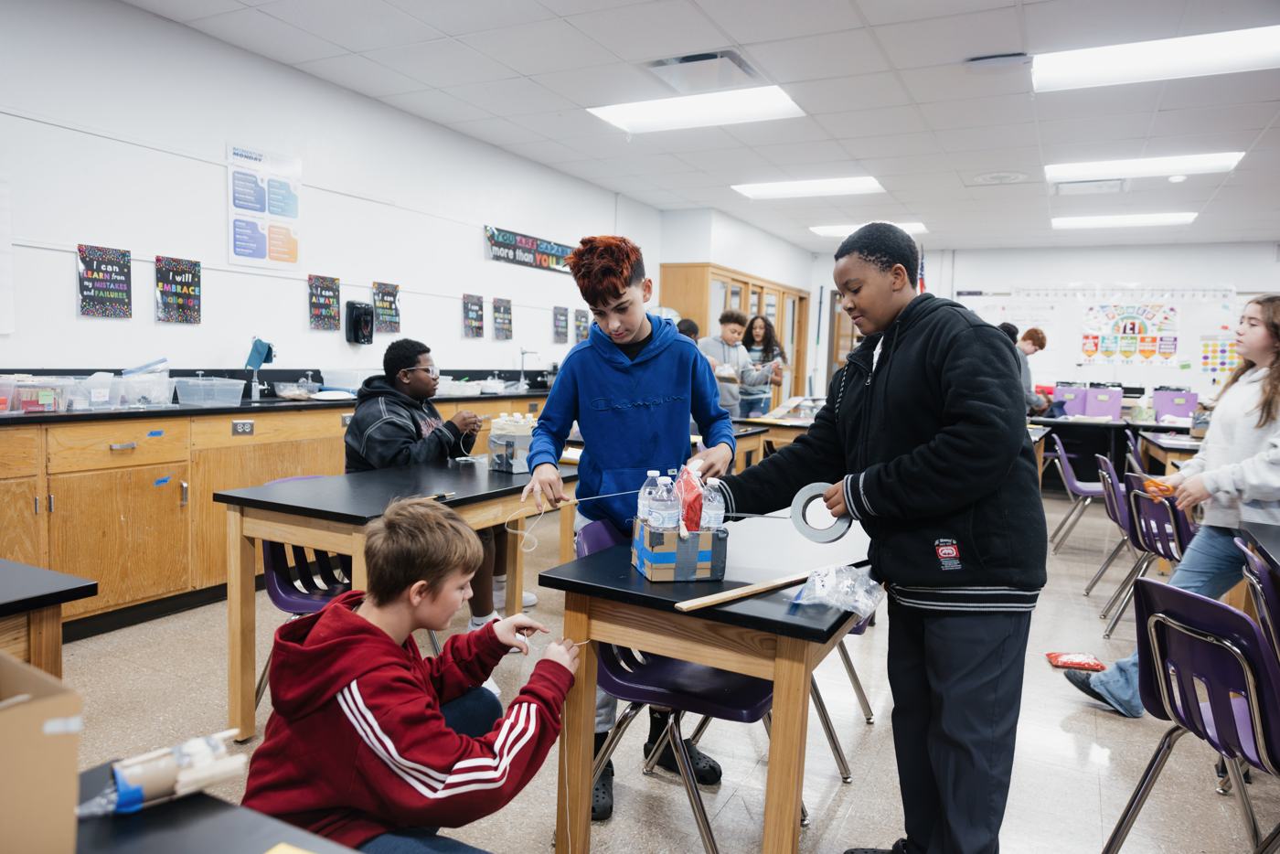 Three Marion students build and tape a box project together at a lab table, measuring string and stacking water bottles.