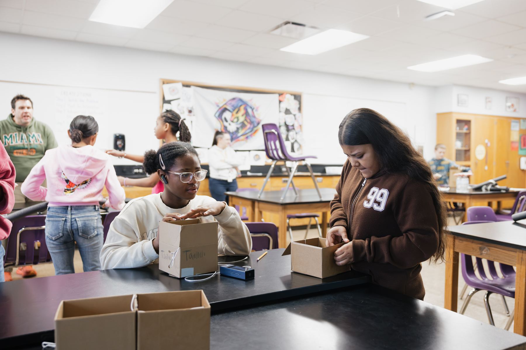 Two Marion students work on cardboard box builds at a lab table during the Exploration Block while classmates and teachers work in the background.